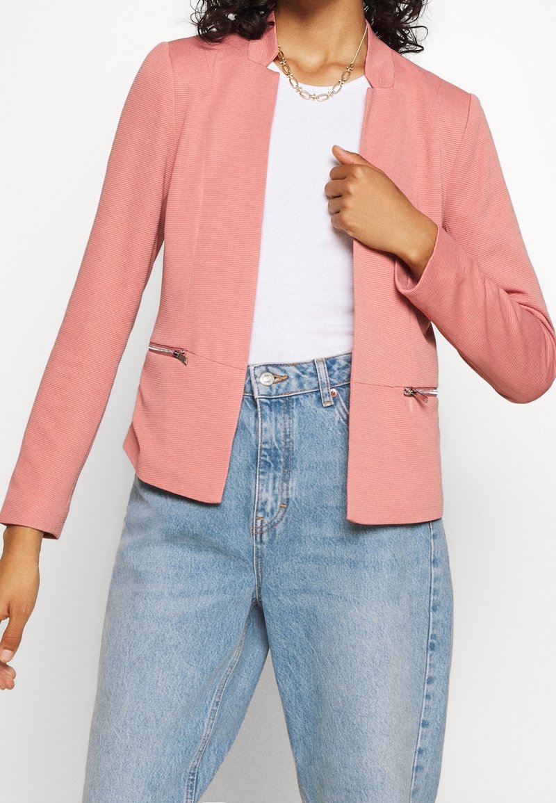 Femme portant un blazer rose clair sur une chemise blanche et un jean taille haute bleu clair, tenant le revers du blazer d'une main.