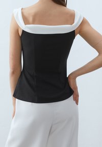 Černý přiléhavý top s bílým skládacím límcem, strukturovaný design, hladká textura a peplum lem, nošený s bílými kalhotami na světlém pozadí.