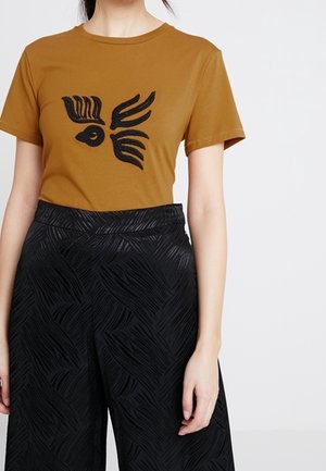 Sininen keltainen T-paita, jossa on musta brodeerattu lintukuva, yhdistettynä mustiin leveisiin housuihin, joissa on teksturoitu aaltokuosi.