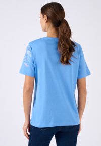 Mujer con una coleta castaña que lleva una camiseta azul de manga corta con un patrón de hojas en la manga izquierda y jeans azul oscuro, vista desde atrás.