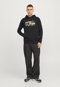 Felpa nera con cappuccio, munita di tasca frontale e stampa grafica in oro e bianco. Abbinata a pantaloni cargo scuri a vestibilità ampia e scarpe sportive.