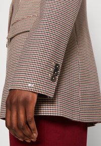 Blazer de patrón de pata de gallo en beige, burdeos y azul marino. Presenta cuatro botones negros en el puño y un diseño de bolsillo elegante.