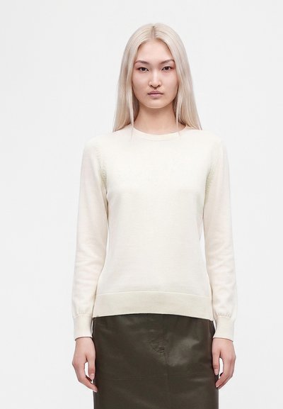 Femme aux longs cheveux blonds portant un pull crème et une jupe vert foncé, debout devant un fond clair uni.