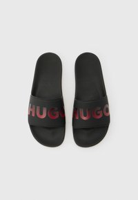 Ζευγάρι μαύρα σανδάλια slip-on με υφασμένα πέλματα και φαρδιές λουρίδες που φέρουν κόκκινες λογότυπες "HUGO" στην κορυφή.