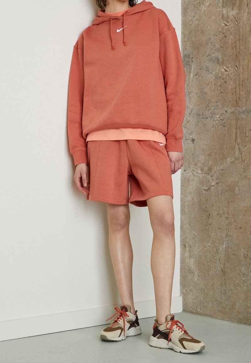 Nike överdimensionerad hoodie i rostfärg med dragsnören, kombinerad med matchande rostfärgade shorts. Vita logotypdetaljer; beige och bruna sneakers fulländar looken.