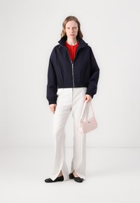 Giacca cropped blu navy con colletto alto, pantaloni bianchi con spacchi laterali e borsa rosa. La modella indossa ballerine nere e una maglia rossa.