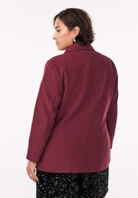 Blazer borgogna con una vestibilità strutturata, maniche lunghe e colletto classico. La parte posteriore presenta un design senza cuciture, abbinato a una gonna nera con paillettes.