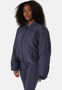 Frau mit lockigem Haar, die eine dunkelblaue, oversized Zip-Jacke und passende Leggings trägt, steht vor einem schlichten, hellen Hintergrund.