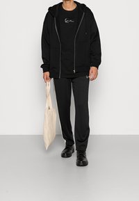 Svart hoodie med dragkedja över en svart t-shirt, kombinerad med svarta byxor med sidostrippar. Håller en ljusbeige tygväska. Enfärgade plagg med avslappnad passform.