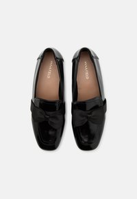 Mocasines de charol negros con un lazo en la parte delantera. Textura suave y brillante, y forma redondeada. Inscrito con el nombre de la marca en el interior.