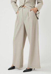 Beige vida byxor med framfållar, hög midja, fickor, ihop med svarta spetsiga kitten heels och en matchande beige blazerkavaj.