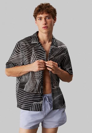 Camicia a maniche corte bianca e nera, con motivi a spirale, colletto aperto e vestibilità comoda, abbinata a pantaloni corti azzurri chiari.