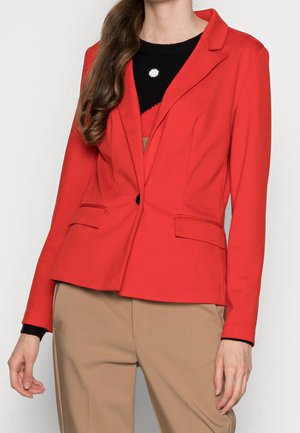 Mujer con una blazer roja ajustada sobre un top negro y rojo, combinada con pantalones beige, de pie frente a un fondo claro y liso.