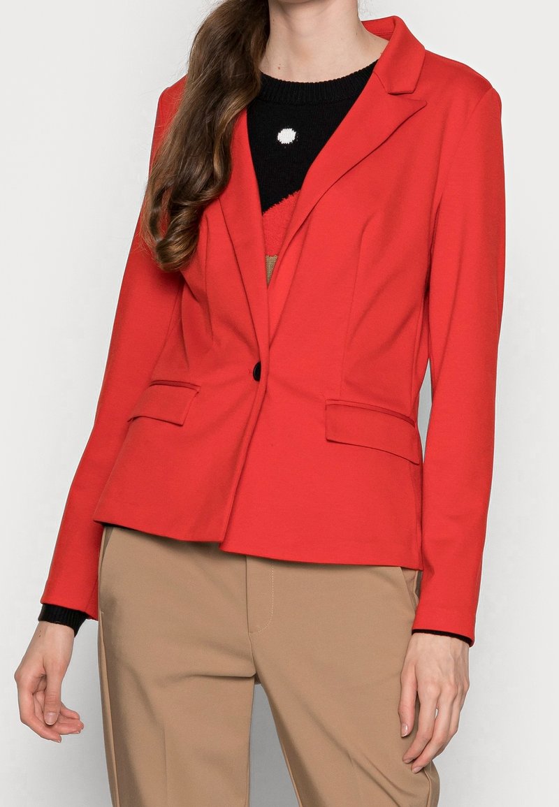 Femme portant un blazer rouge ajusté sur un haut noir et rouge, associé à un pantalon beige, debout devant un fond uni clair.