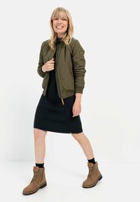 Olivgrüner Bomberjacke mit gerippten Bündchen, getragen über einem schwarzen gerippten Kleid, kombiniert mit braunen Schnürstiefeln. Glattes Material und sportliches Design.