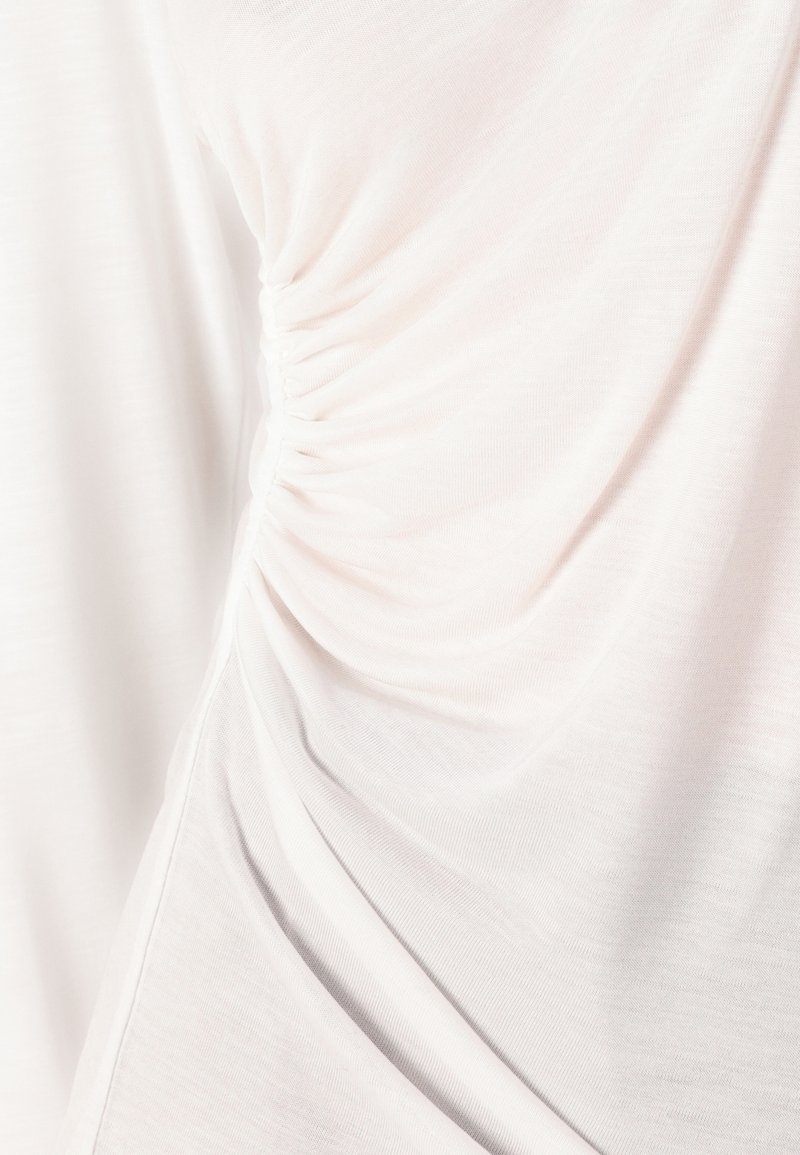 Jasnoróżowa bluzka z miękkiego, teksturowanego materiału, z marszczonymi detalami po bokach, co tworzy efekt drapowania.