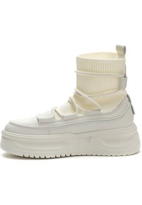 Zapatillas blancas con parte superior suave, cuello de calcetín acanalado y suela gruesa que presenta patrones texturizados y detalles elásticos.