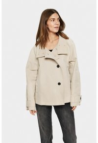Beige dvouřadý kabát s velkým límcem a výraznými knoflíky. Má široké rukávy a volný střih, je vyroben z hladkého materiálu.