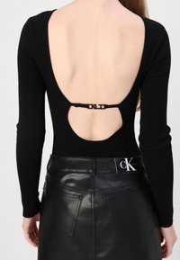 Black ribbed top with long sleeves and a deep back cutout, secured by a black strap. Paired with a black leather skirt featuring pockets.