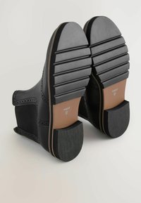 Mustad nahkkingad, millel on tekstureeritud kummist tallad, dekoratiivsed brogue-detailid ja sile viimistlus. Kontsad on massiivsed ja unikaalse soonilise mustriga.