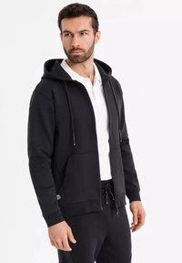 Sort hættejakke med lynlås og snøre, blødt stof, kængurulommer, båret over en hvid polo t-shirt, kombineret med sorte sweatpants.