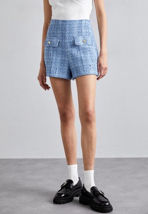 Frau trägt blaue, strukturierte Shorts mit hohem Bund und vorderen Klappentaschen, weiße Socken, schwarze Loafers und ein weißes ärmelloses Top.