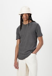 Black and white striped t-shirt with a round neckline, short sleeves, and a curved hem, paired with cream-colored wide-leg pants and a beige bucket hat.