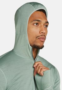 Giacca a zip verde chiaro con cappuccio, caratterizzata da dettagli in texture, progettata con fori per i pollici, dotata di una zip completa e di un cappuccio aderente.
