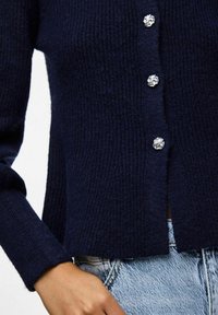 Zila adīta kardigāna ar faktūru, kam ir pieci dekoratīvi sudraba pogas priekšpusē un īsa apakša.
