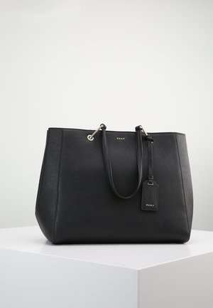 Czarna skórzana torba typu tote DKNY z podwójnymi uchwytami i przymocowaną oznaczoną metką bagażową na białej powierzchni.