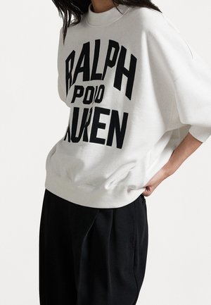 Vrouw draagt een witte oversized trui met zwarte tekst "RALPH POLO LAUREN", gecombineerd met losse zwarte broek, handen in de zakken.