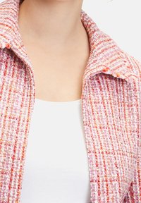 Różowo-pomarańczowa kurtka z tweedu o teksturowanej powierzchni, z szerokim kołnierzem i otwartym przodem. Brak widocznych zapięć.