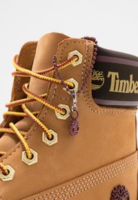 Gros plan d'une botte Timberland couleur fauve avec des lacets jaunes et rouges, des œillets dorés et une étiquette logo violette suspendue à une chaîne de perles.