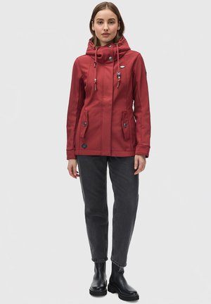 Veste imperméable rouge avec un col haut, capuche avec cordon de serrage, poches avant et boutons-pression. Associée à un jean foncé et des bottines noires.