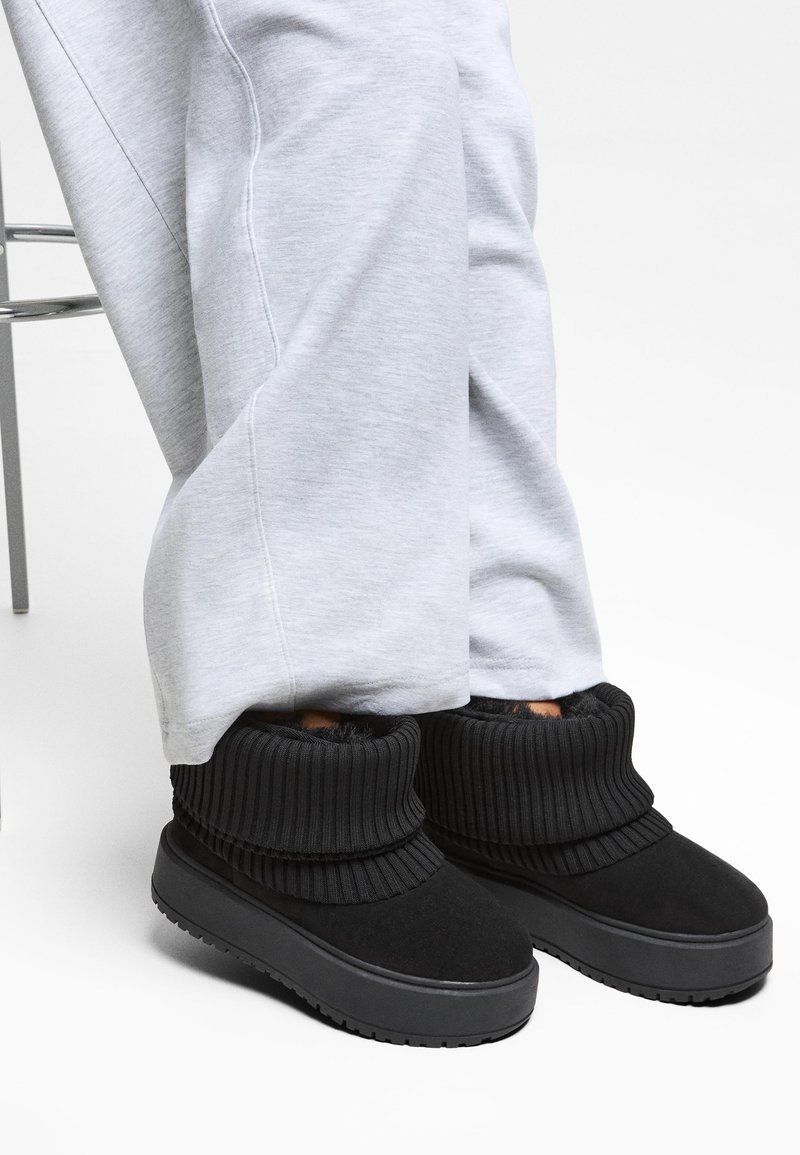 Personne portant des bottines noires avec des revers côtelés retournés, associées à un pantalon ample gris clair, debout à côté d’une chaise en métal.