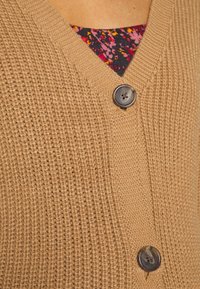 Cardigan marrone lavorato a maglia con scollatura a V, caratterizzato da due bottoni e un tessuto testurizzato. All'interno, è parzialmente visibile una maglietta con motivo floreale.