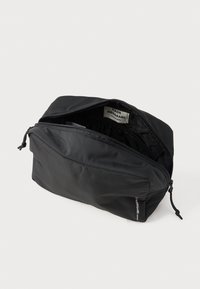 Crna torba na zatvarač od najlonske tkanine, s prostranim unutarnjim prostorom i istaknutom oznakom marke unutra. Glatka tekstura s suptilnim sjajem.