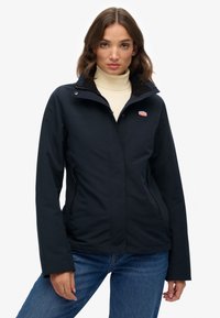 Marineblauwe waterdichte jas met een hoge kraag, twee met rits afgesloten zakken en een logopatch op de borst. Bevat lange mouwen en een gladde textuur.