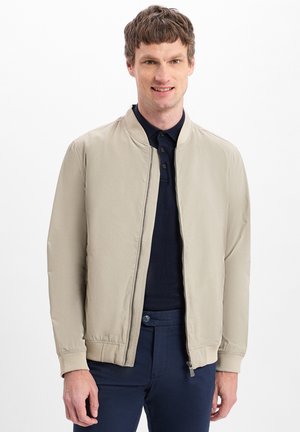 Beige Jacke im Bomberdesign, mit geripptem Kragen und Bündchen. Mit Reißverschluss vorn und Seitentaschen, getragen über einem marineblauen Poloshirt.