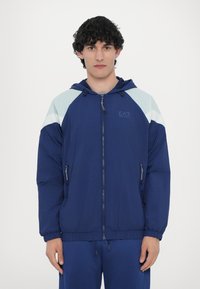 Navy zip-up jacket with a hood, featuring light blue accents on shoulders, elastic cuffs, and side pockets. Smooth, lightweight material.