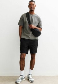 Grå kortärmad t-shirt, svarta cargoshorts, svart axelväska, vita ankelsockor och grå sneakers med detaljer. Takbelysning, neutral bakgrund.