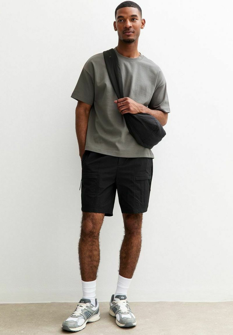 Grå kortärmad t-shirt, svarta cargoshorts, svart axelväska, vita ankelsockor och grå sneakers med detaljer. Takbelysning, neutral bakgrund.