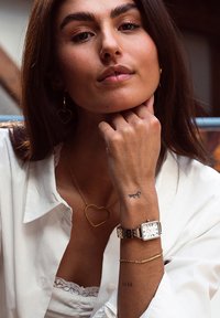 Montre dorée avec un cadran rectangulaire, un fond blanc et de grands marqueurs. Comprend des colliers en or superposés et des boucles d'oreilles en forme de cœur.