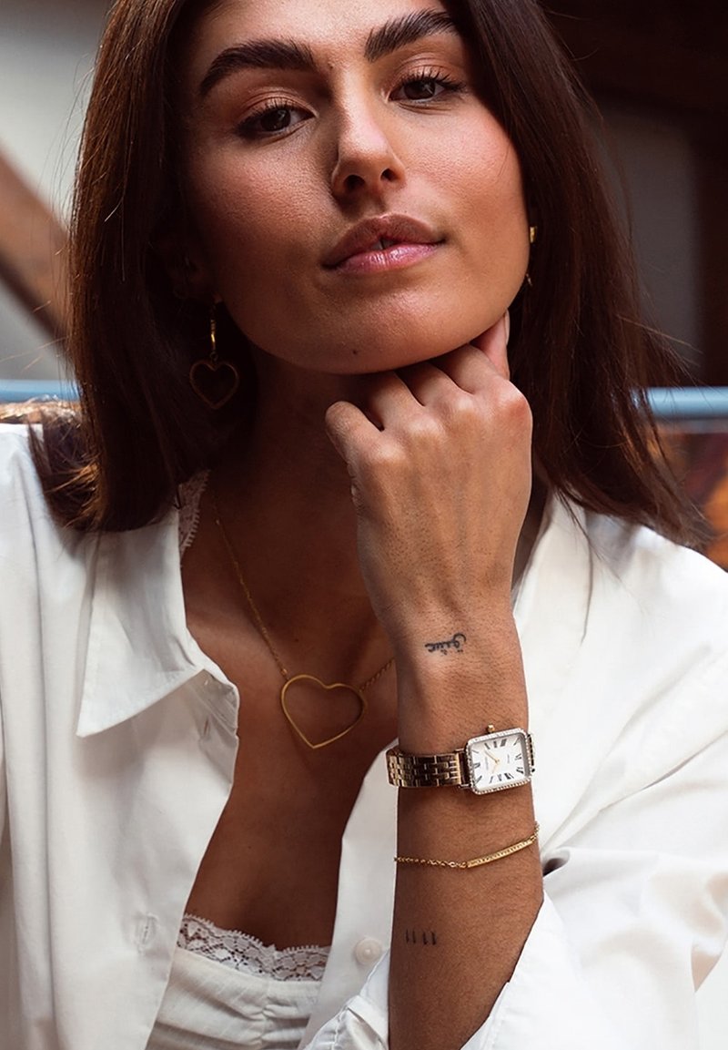 Montre dorée avec un cadran rectangulaire, un fond blanc et de grands marqueurs. Comprend des colliers en or superposés et des boucles d'oreilles en forme de cœur.
