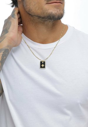 Mann in weißem T-Shirt trägt eine goldene Kettenhalskette mit einem schwarzen, rechteckigen Anhänger, der einen goldenen Stern zeigt; eine Hand berührt den Hals und zeigt einen tätowierten Arm.
