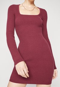 Ribbad stickad klänning i bourgogne med långa ärmar, fyrkantig halsringning och figurnära silhuett. Slät textur med subtil stretch.