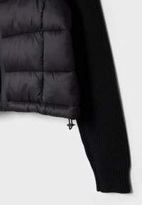 Doudoune noire avec une texture matelassée d'un côté et un tissu en tricot côtelé de l'autre, dotée d'une fermeture éclair et d'un cordon de serrage au niveau de l'ourlet.