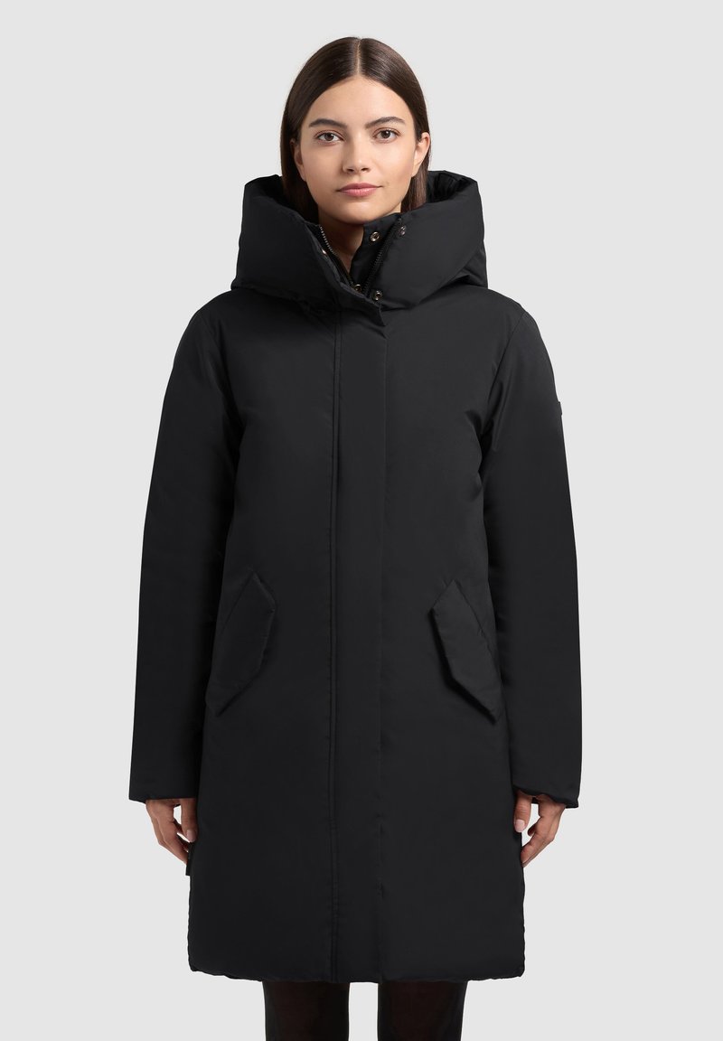 Sort polstret frakke med høj krave, lynlås lukning, sidelommer og lange ærmer. Glat tekstur og enkelt design.