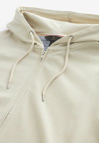 Khaki mikina s texturou, s kapucí na stažení a stříbrnými kovovými doplňky. Vnitřek má geometricky vzorovanou podšívku.