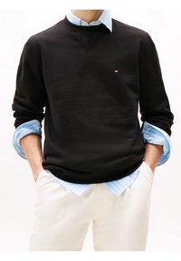 Felpa nera con colletto rotondo, polsini a costine e un piccolo logo. Indossata sopra una camicia a righe blu chiaro, abbinata a pantaloni bianco sporco.